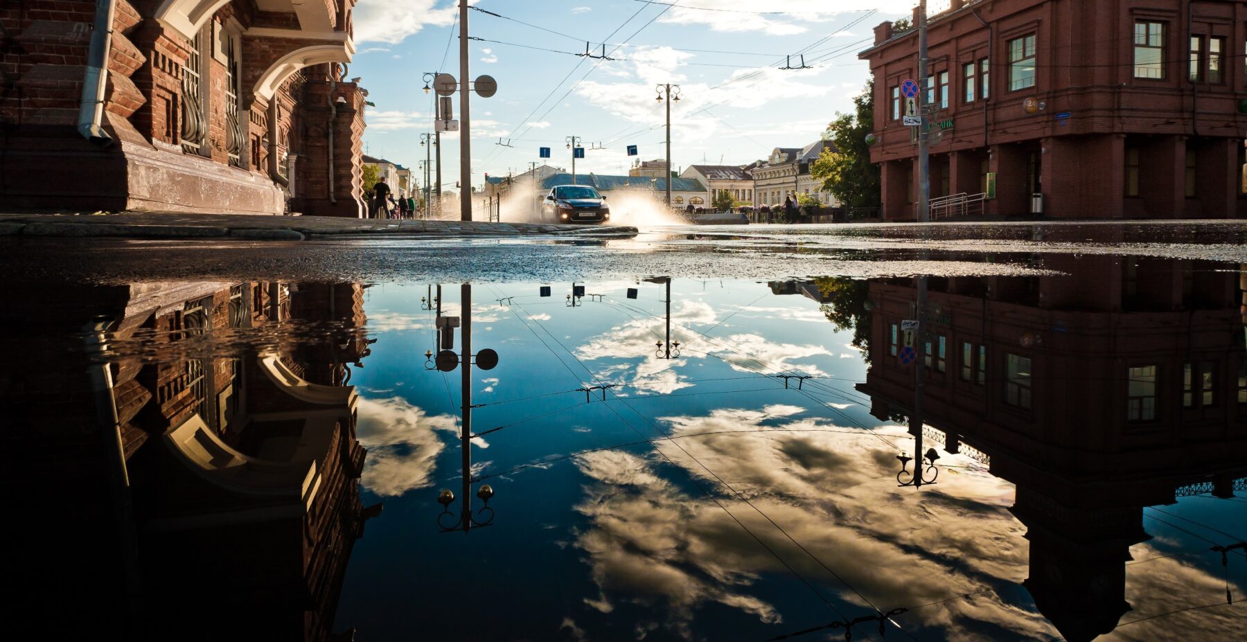 Urban street with reflective puddle after rain, showing buildings and sky; a car drives through, highlighting flood-resilient design needs.