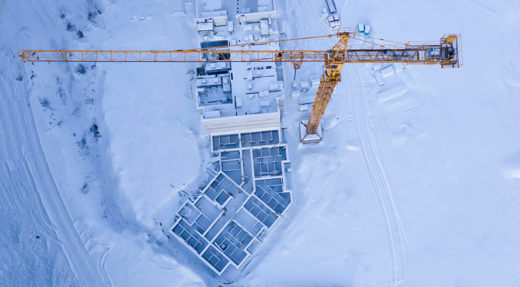 Aerial view of a snow-covered construction site with a crane and foundations, illustrating spring construction planning and material delivery scheduling.